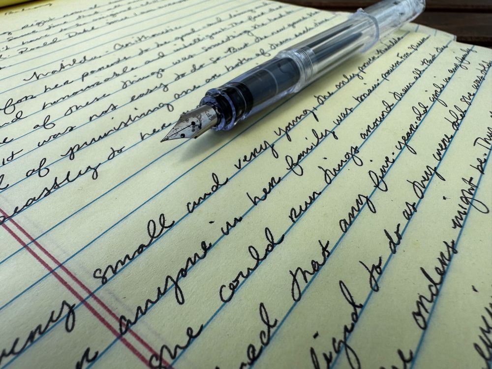 A close-up image of a fountain pen resting on a yellow legal pad filled with neat, cursive handwriting in black ink. The pen is transparent with visible ink inside, and the writing lines are blue with a red vertical margin line on the left. The perspective is angled, emphasizing the elegant script and the nib of the pen.