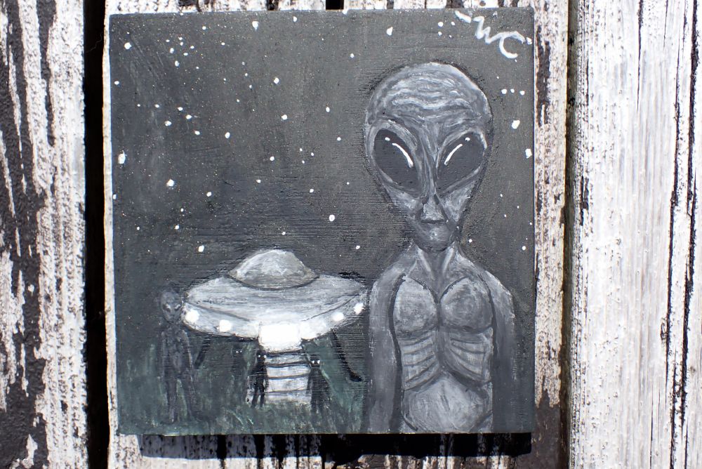 A painting of a grey alien and a UFO. The alien is to the right and staring at you. The UFO is behind it and to the left, and has the silhouettes of other aliens around it. 