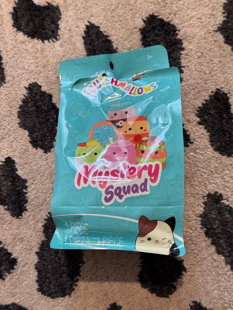 an opened blue mystery squad squishmallow bag laying atop a black spotted rug 