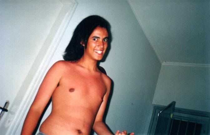 Álvaro (18? 19?) com cabelo preto comprido alisado, pele morena, de torso nu, com um sorriso maníaco