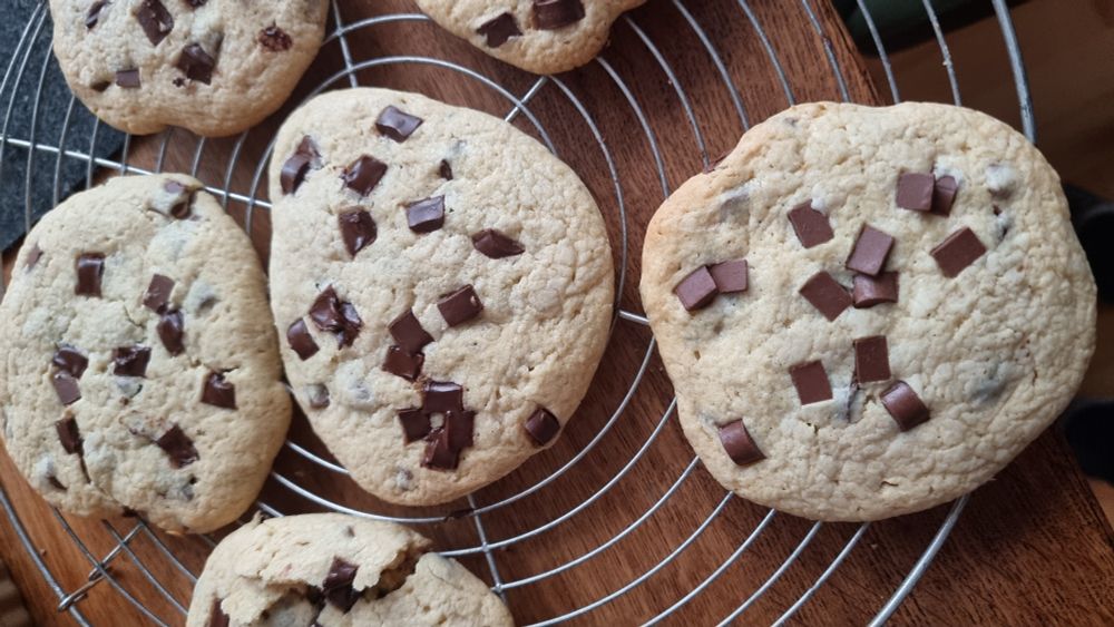 Große, helle Chocolate-Chip-Cookies liegen auf einem runden Metall-Kuchengitter; die weichen Kekse sind ungleichmäßig geformt und mit dunklen Schokostückchen bestreut, der Hintergrund besteht aus einer Holzoberfläche.

