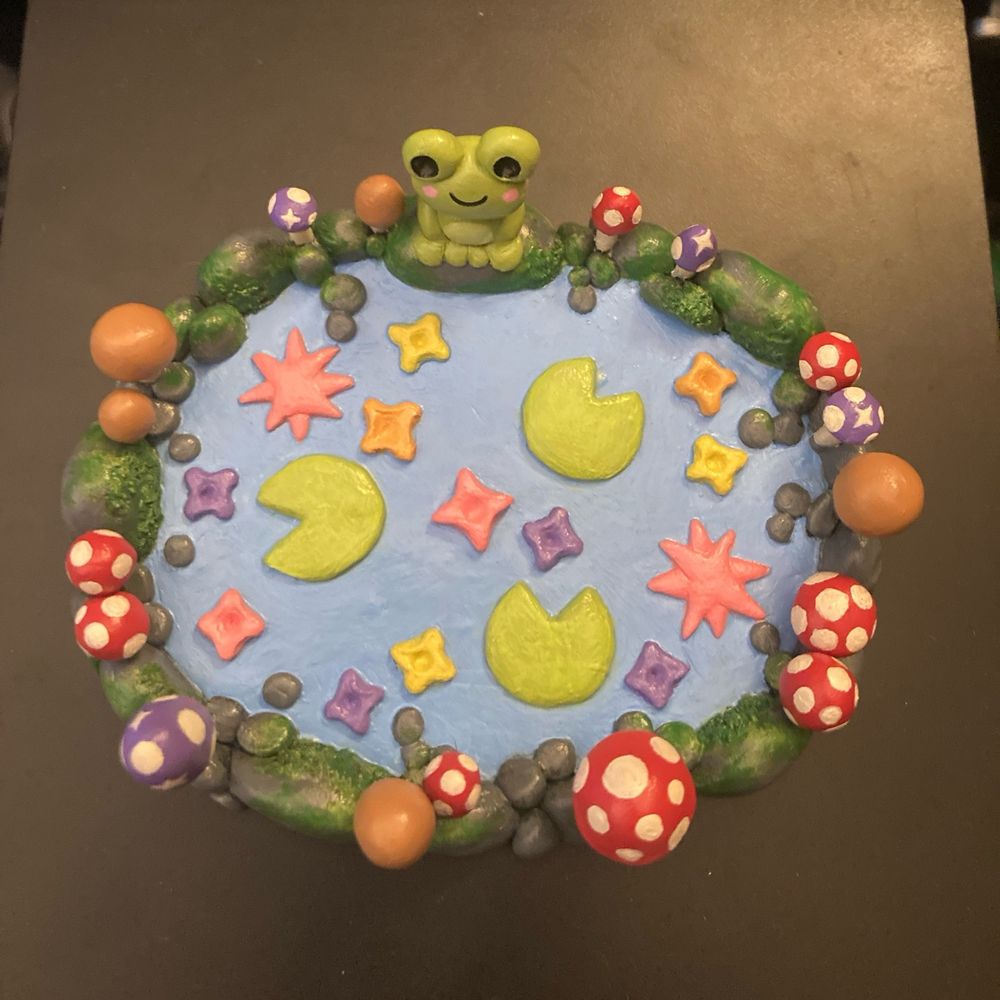(an aerial view of the same trinket dish.) an oval shaped trinket dish made from polymer clay and painted. the dish is a small pond with a border of stones. a cartoonish frog with a smile and pink blushies sits atop one of the stones. there are brown, red, and purple mushrooms around the border, and some moss is growing on the rocks. inside the pond, floating on the surface of the "water" are bright green lily pads and flowers painted pink, purple, orange, and yellow.