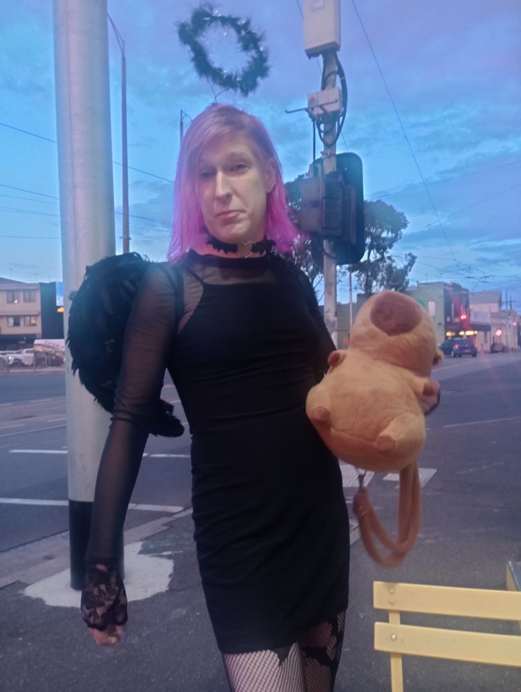 White trans woman with purple pinky hair wearing short black dress black choke and with black halo and black feather wings. Also holding a bag in the shape of a capybara with an orange on its head. What you can't see is the kick arse boots but they're implied 