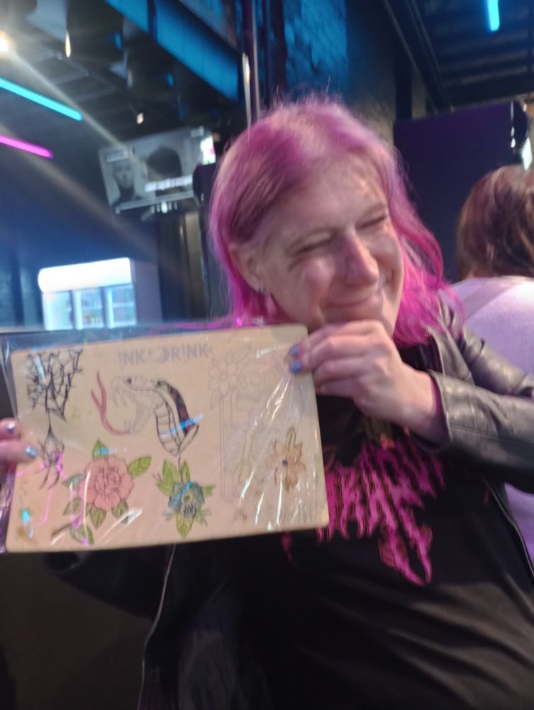 Purple haired trans woman holding up a piece of silicon with faux tattoos on it. From top left spider, snake, skeleton in ☕ b, two flowers. Only the rose looks semi decent