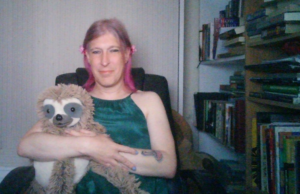 MEGA Clocky middle-aged trans woman with pink hair sitting in a home office wearing a too-tight green dress that leaves her shoulders bare. Here hair is held back in pink flower clips. She's holding a stuffed sloth. It can just be seen she has a tattoo of a pink and blue amanita mushroom on her left arm. She has an awkward smile on her face, perhaps as if she's STILL not used to selfies and STILL really uncomfortable pretending anyone should ever see this shit.