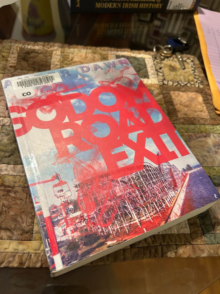A copy of Sodom Road Exit, by Amber Dawn, is displayed on an earth-toned placemat. The text is red, tightly kerned and sans serif, and there is a garish photo of a roller coaster under a blue, cloud-studded sky.