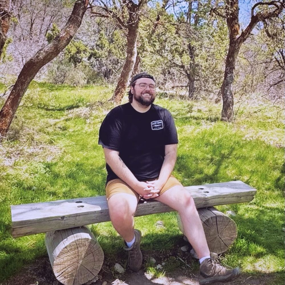 A picture of me, smiling and enjoying the sun, on a bench along a hiking trail 