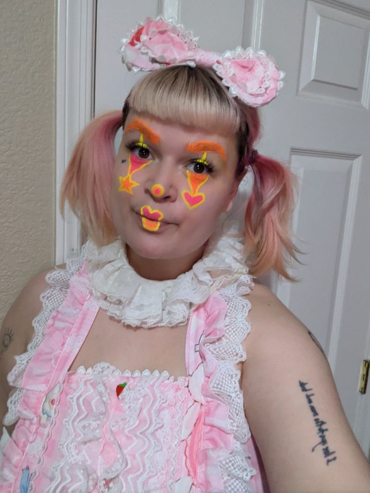 a selfie wearing orange and pink clown makeup and a pink dress with a clown collar
