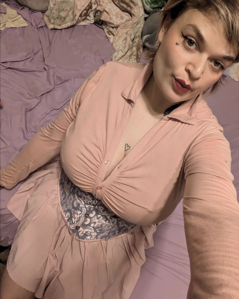 Lush sitting down with a pink long sleeve blouse, blue and pink corset, and a pink skirt.