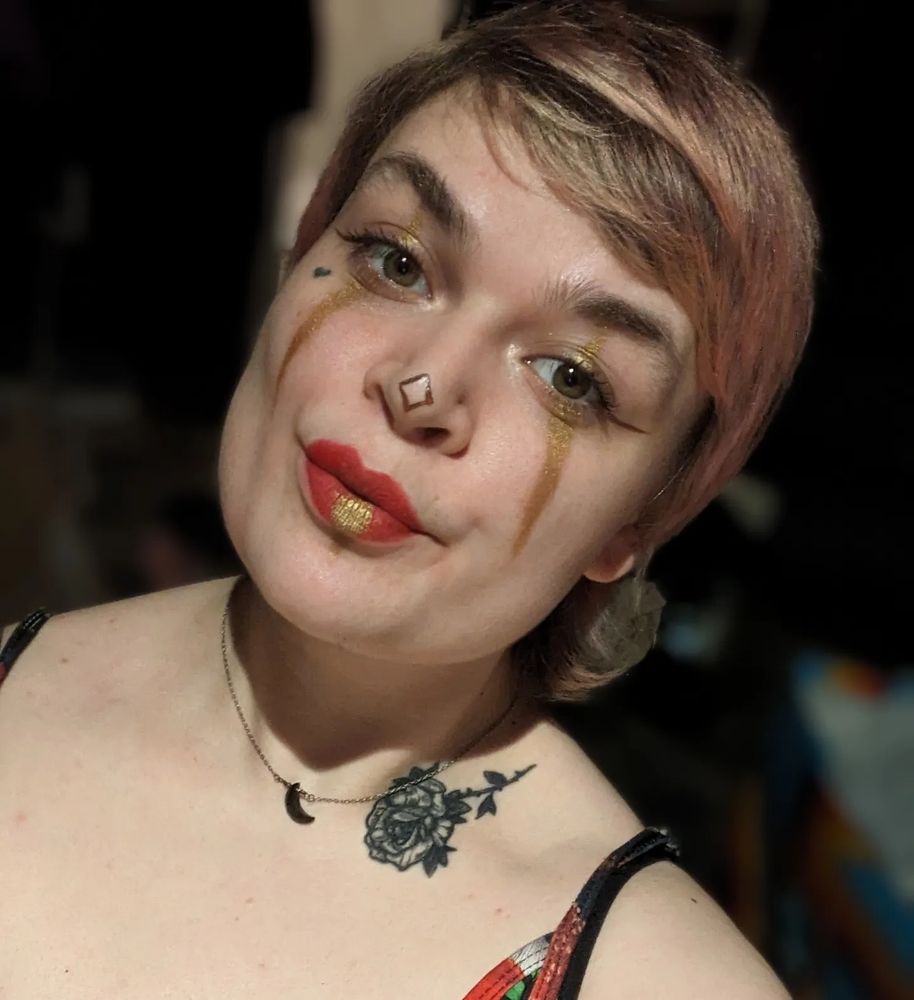 lush with jester makeup on. gold eye makeup, red and gold lips, a brown outline of a diamond on the nose, brown horizontal eyeliner wings)