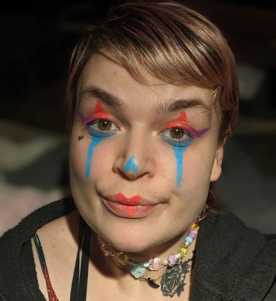Lush in clown makeup (coral, blue, and purple eye makeup, upside down blue heart on the nose, and a coral heart on the lips)