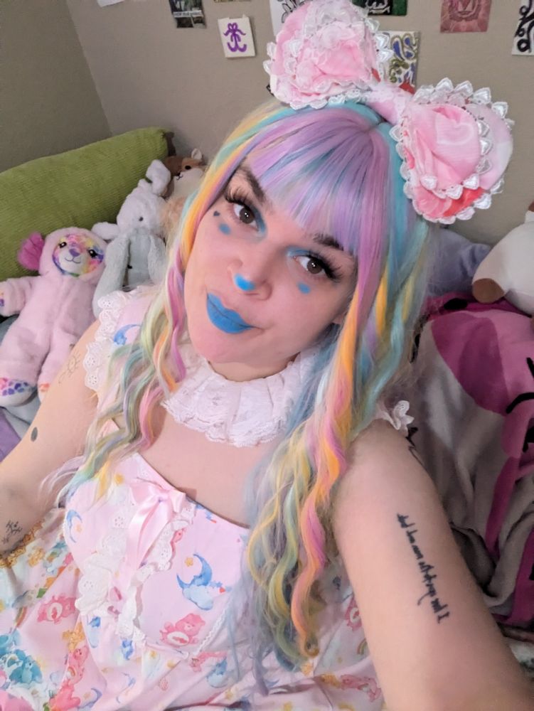 Lush in blue clown makeup with a frilly white jester collar and a pink dress with care bears on it, they're wearing a large pink bow in their multicolor pastel hair