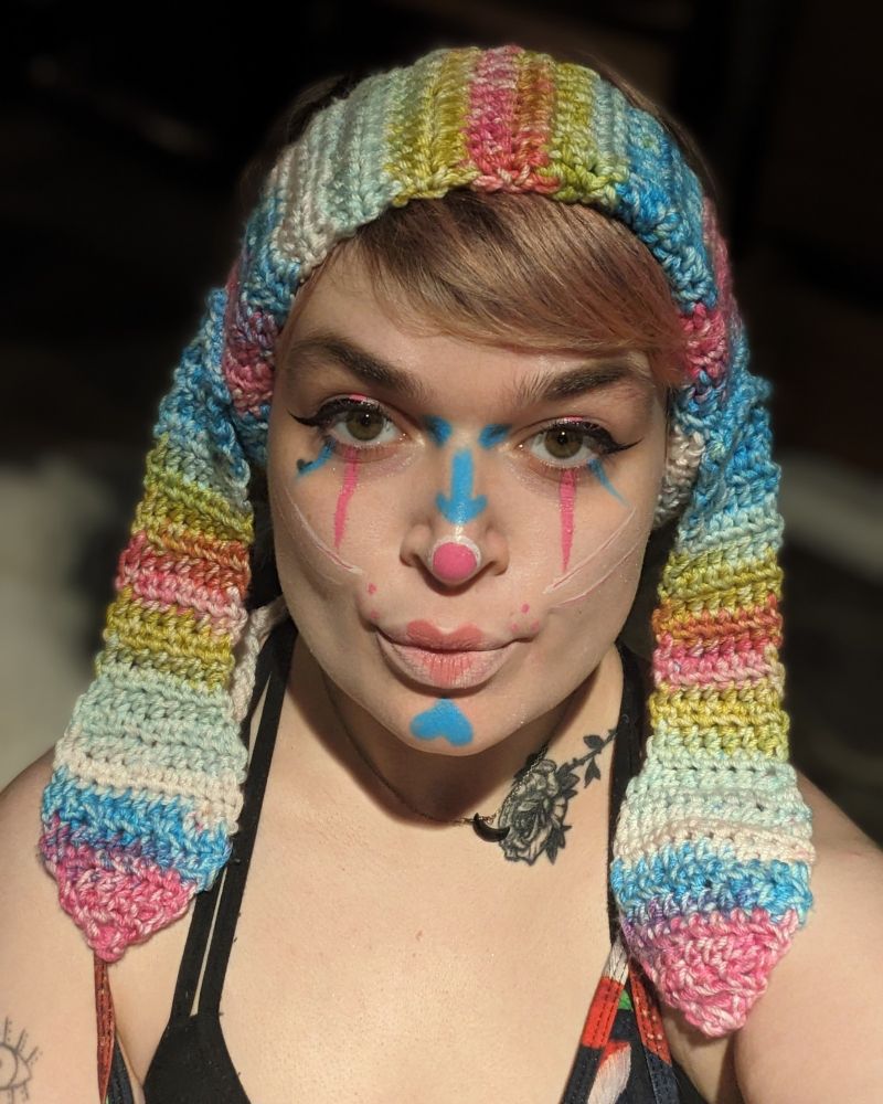 Lush being a bunny jester (wearing a multicolor bunny ear crochet headband, with blue and pink clown makeup and white whiskers)