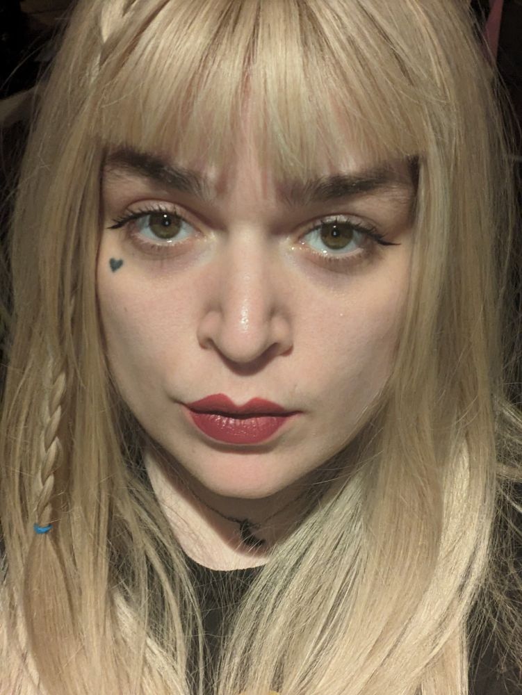 lush in a blonde wig with bangs and red lipstick with downturned eyeliner wings