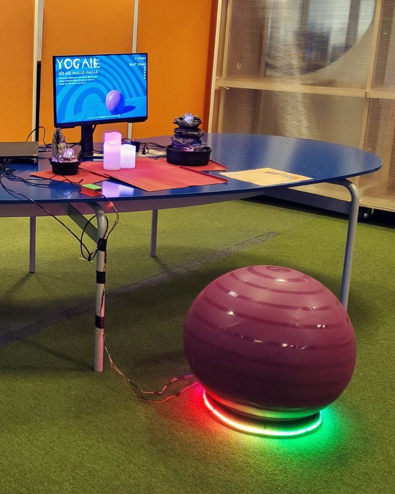 A yoga ball standing in front of a table with a game screen and some decorations such as led candles and led mini-fountains. The yoga ball is put above a bike wheel with an LED strip.