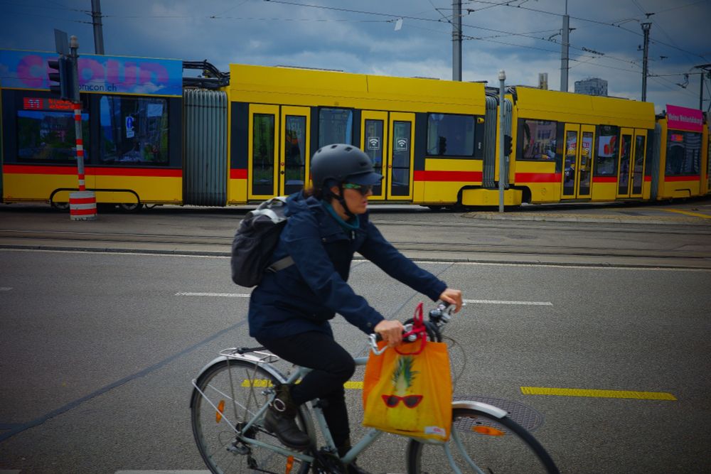 Streetfotografie in Farbe mit einer Ricoh GRIIIx. Im Vordergrund mittig eine Person mit Fahrrad. Am Lenker hängt ein Plastiksackerl in gelber Farbe. Dahinter eine Tram der Baselland Transport AG (kurz BLT) in den Farben Gelb und Rot.