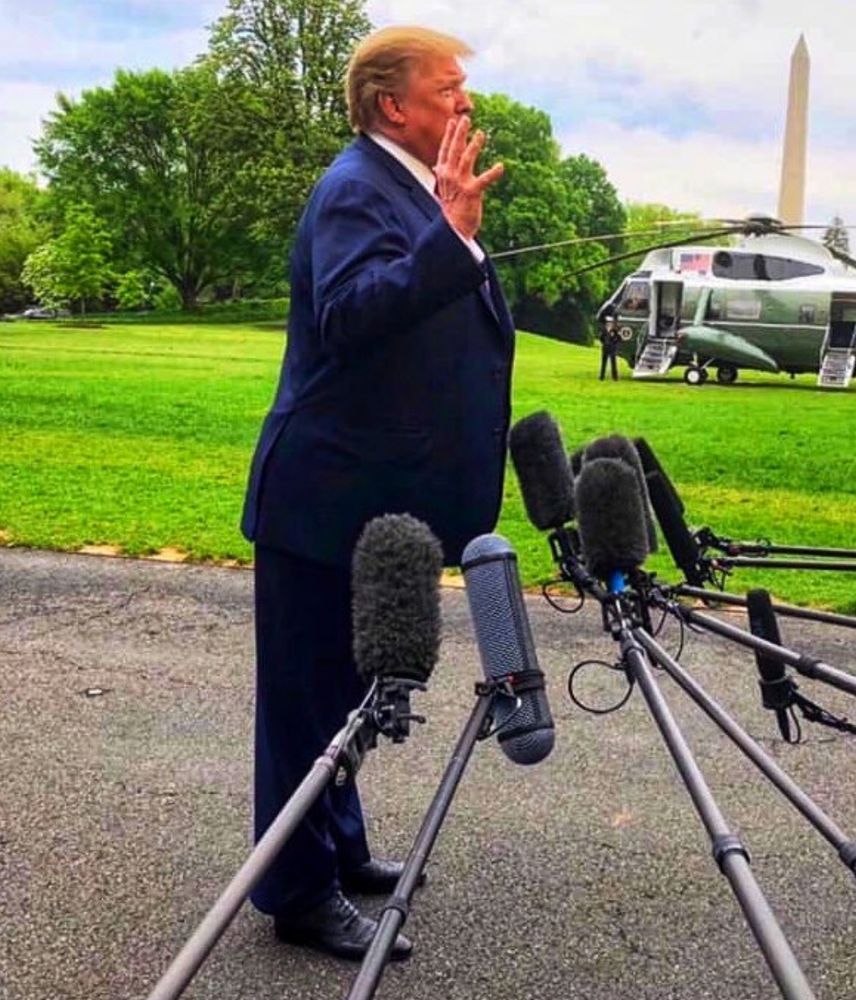 Trump in 1st term. The image shows his profile. He is leaning extremely far forward. A bunch of microphones are held by offscreen workers at about waist height. Trump's hands are in the air as if he is talking with them. A helicopter is in the background.