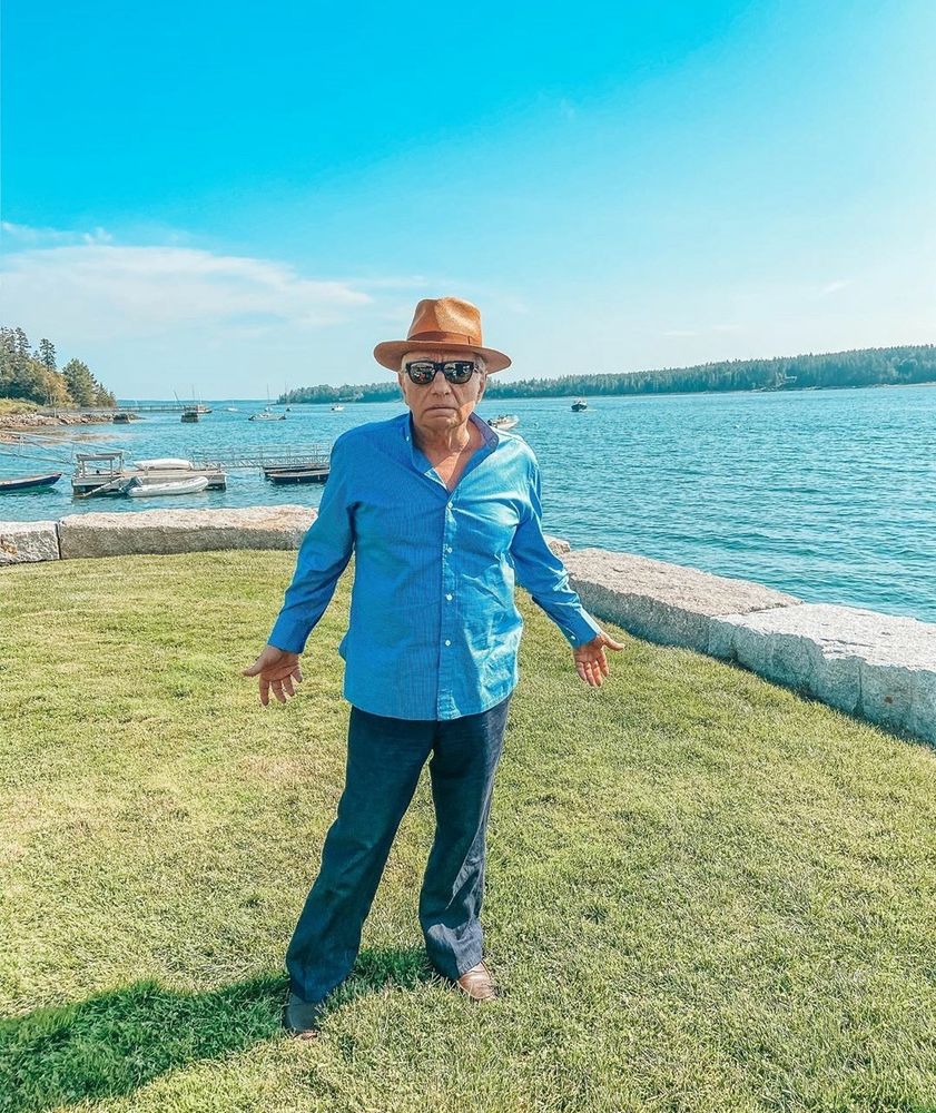 Martin Scorsese wearing a bright blue button down shirt, sunglasses, and brown fedora standing on green grass with arms outstretched by the water