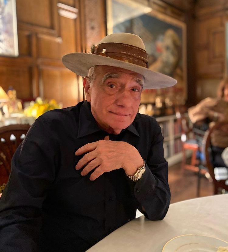 Martin Scorsese wearing a black shirt and large, wide brimmed hat sitting at a table posing for the camera 