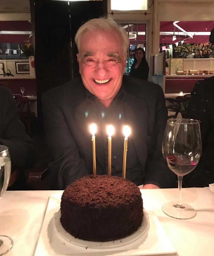 Martin Scorsese grinning behind a round, chocolate cake with three candles and a glass of red wine nearby
