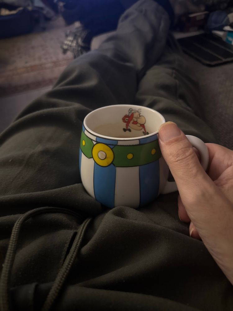 Eine bauchige Kaffeetasse mit blau-weißen Streifen außen und einem Obelix-Kopf auf der Innenseite, man sieht einen Teil der haltenden Hand und die Beine in Jogginghose, auf denen die Tasse steht. 
