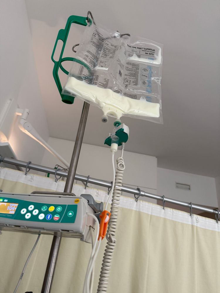 Im Krankenzimmer: An einem Infusionsständer hängt ein Beutel mit intravenösem Nahrungsbrei. Im Vordergrund (auch an dem Ständer) ein elektronisches Dosiergerät