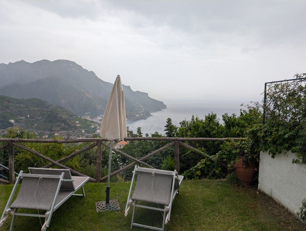 A tall grassy mountain in the distance with a village down below and the Mediterranean Sea to the right. Lawn chairs and an umbrella are in the foreground. 