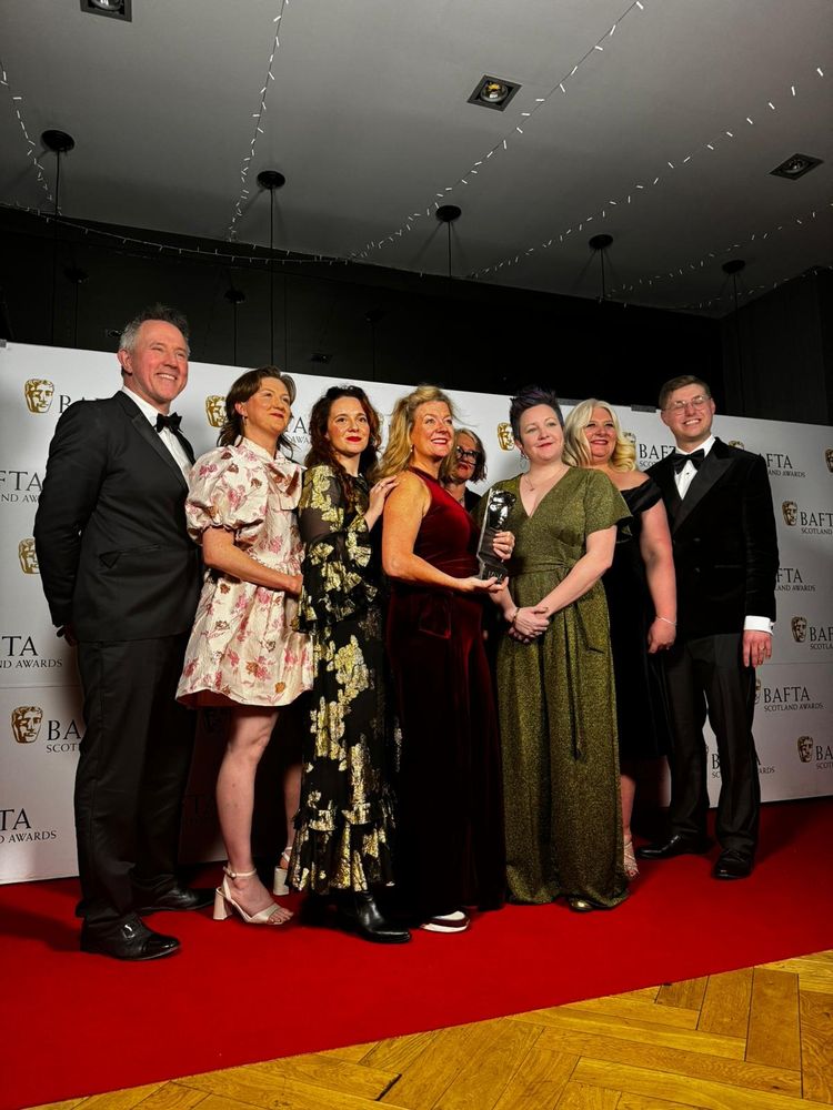 A group of people dressed in formal evening wear posing for an official photographer holding a BAFTA award