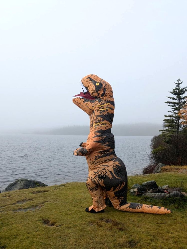 Jeff inside an orange inflatable T-Rex costume. Standing in front of a pond and looking at the sky.