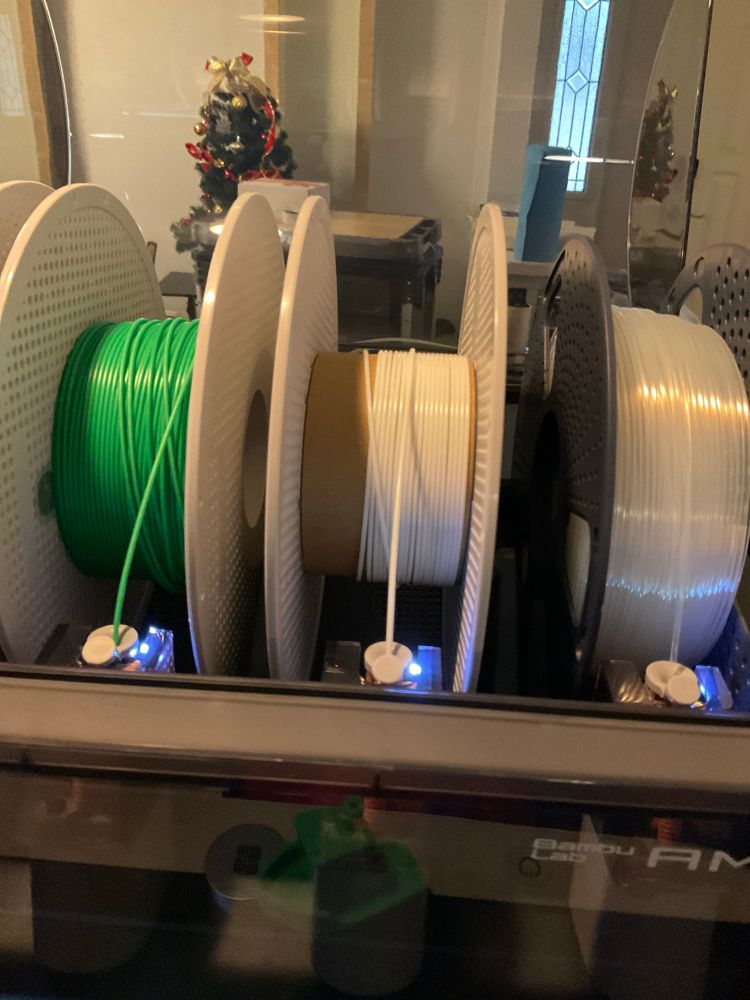 3D printing filament rolls feeding into a printer, the roll of green on the left appearing to be about 10% from empty, the translucent on the right looking mostly full, and the white in the center showing bare spool and maybe 0.5 to 1% from empty. 