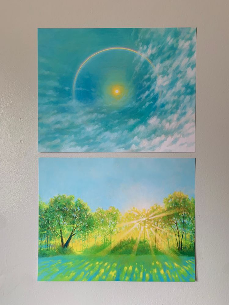 two rectangular prints against a gray background. Top print depicts a blue sky with warping white clouds, and a bright yellow sun surrounded by a rainbow. Bottom print depicts a green landscape of a tree line with the sun bursting through it, casting dappled shadows on the grass below.