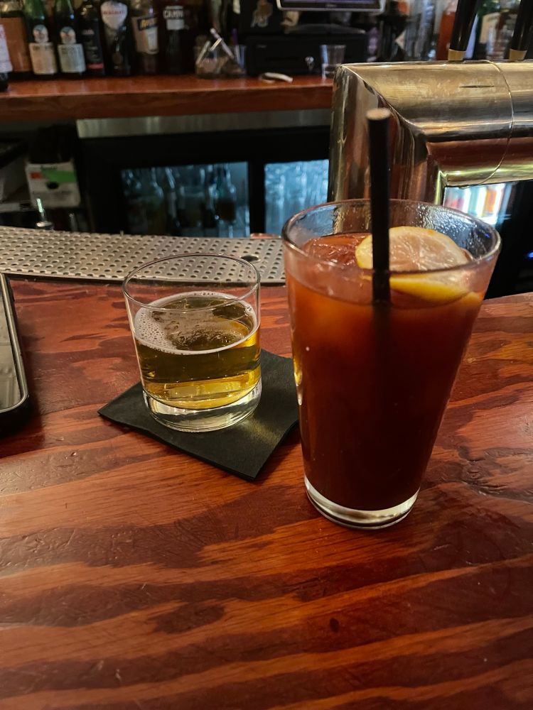 A short glass of beer on a black napkin and a tall glass of mezcal Bloody Mary on a wooden bar top