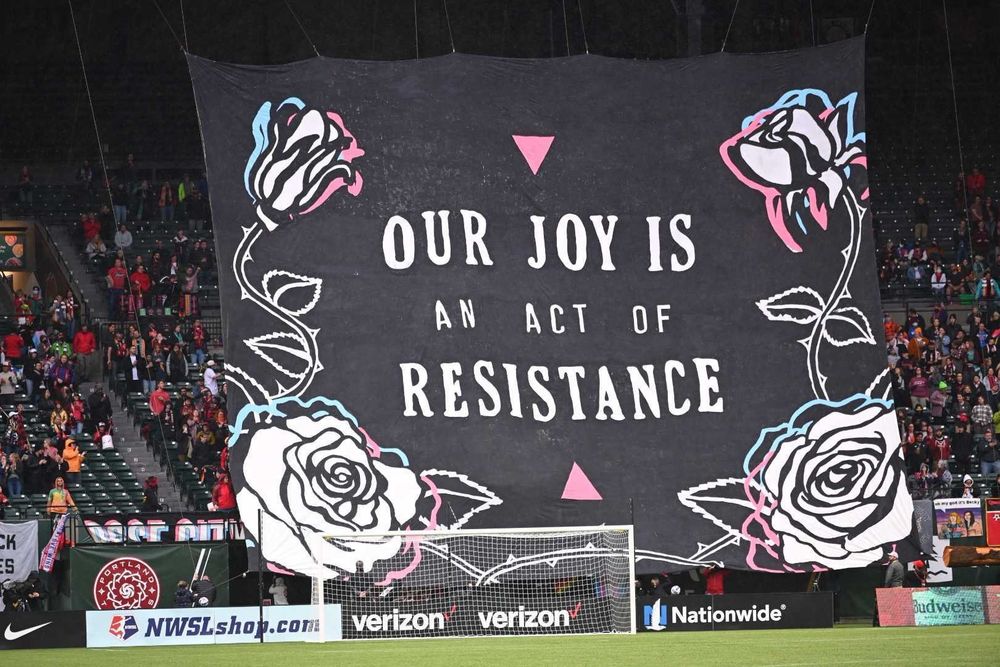 A Rose City Riveters tifo in Providence Park. The design is white on black with echoes of trans pride colors. The text reads: Our joy is an act of resistance. 