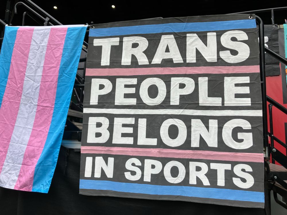 Rail banner next to a trans pride flag. The rail banner reads: trans people belong in sports 