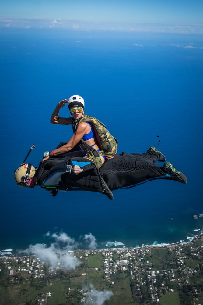 Photo of a “wingsuit rodeo” over Arecibo, Puerto Rico.  In a wingsuit rodeo, a skydiver (using typical skydiving gear) rides on the back of a wingsuited skydiver.  