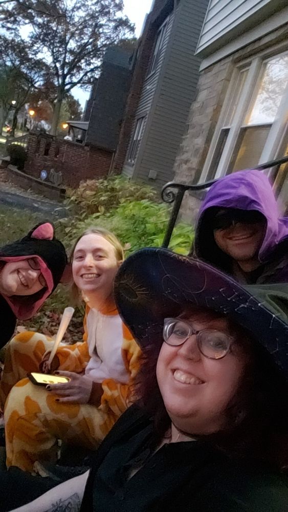 Four people sit on steps getting ready to pass out candy. One is wearing a black cat shawl, one is wearing a giraffe kigurumi, one in black with a witch hat, and one in a mask and a purple cloak.