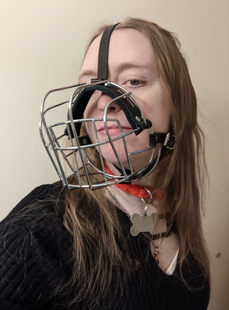 A white woman with long blonde hair stands in front of a white wall.
She is wearing a metal dog muzzle strapped to her face as well as a red collar with a bone shaped tag.
She is wearing a black top with a chest window held together by a clasp.