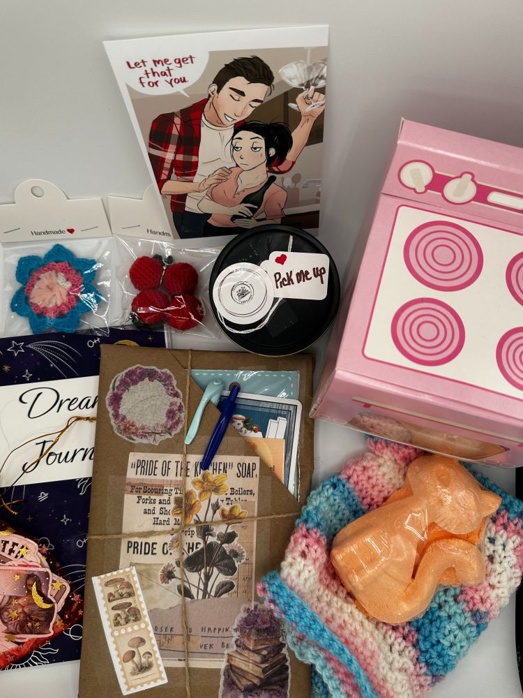 picture of the items available in a cozy book box. Pin, pen, highlighter, book, journal, bookish stickers, hat, bath bomb, fuzzy socks, character art. 