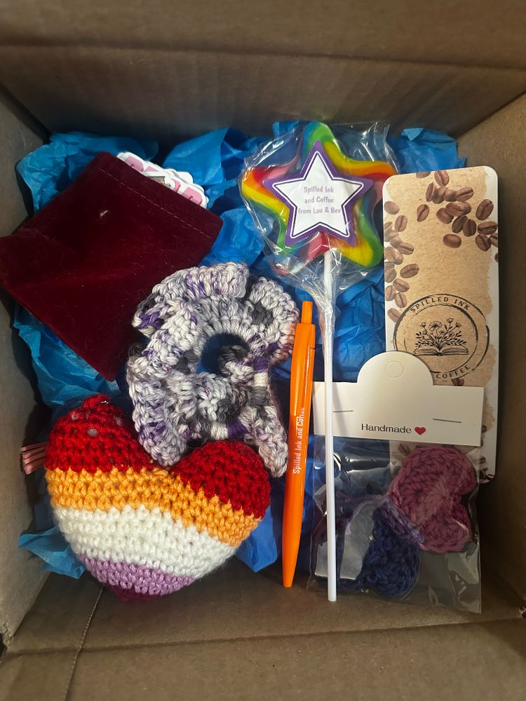 Box filled with an orange pen, large rainbow lolly, bi pride butterfly pin, ace pride scrunchie, lesbian pride heart, small bag of bookish stickers, spilled ink and coffee bookmark. 