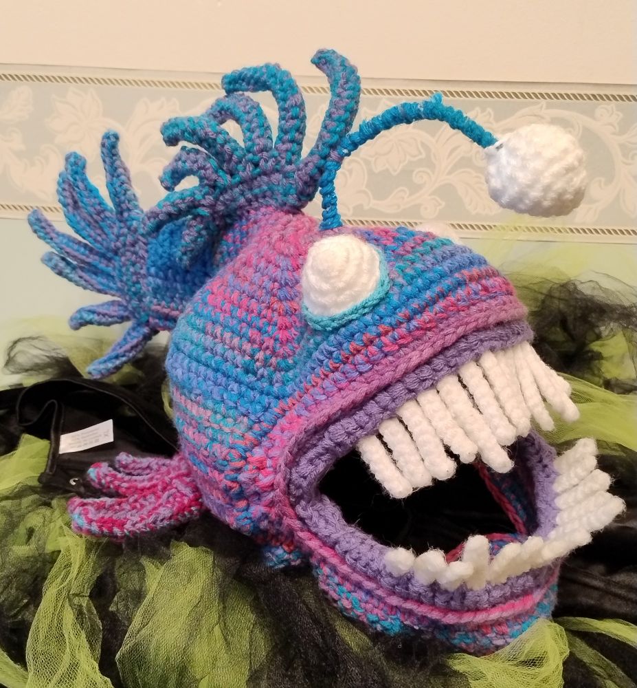 Crocheted angler fish hat in blue pink and purple, resting on a green and black striped tutu. Hey all, despite not having interacted with the dead bird since 2020, I have now deleted my account there. Hopefully this place will be lovely. Hope you enjoy the photo of the hat that I made last month! #crochet #🧶