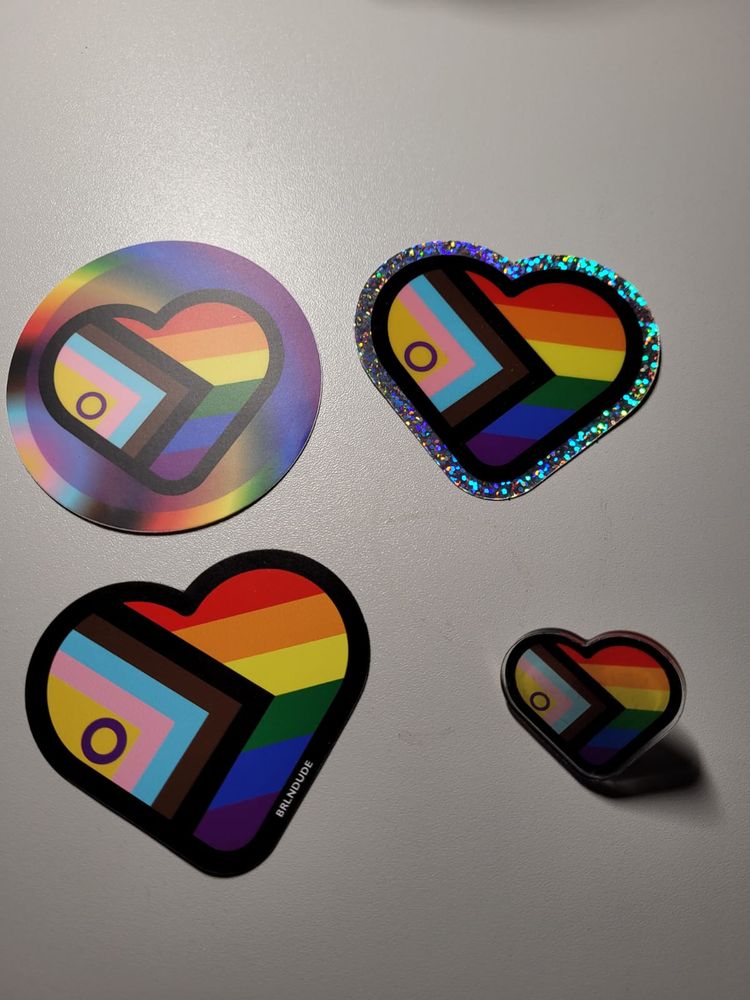 Oben 2 Aufkleber in den Farben der ProgressPride Flag. Der linke ist Rund und mit der Flagge in Herzform in der Mitte. Der Rechte ist ein Herz in den Farben der ProgressPrideFlag mit einem Rand aus Glitzer. 
Unten Links ein flaches Herz in den Farben der ProgressPrideFlag und unten links ein Pin in Herzform in den Farben der ProgressPrideFlag