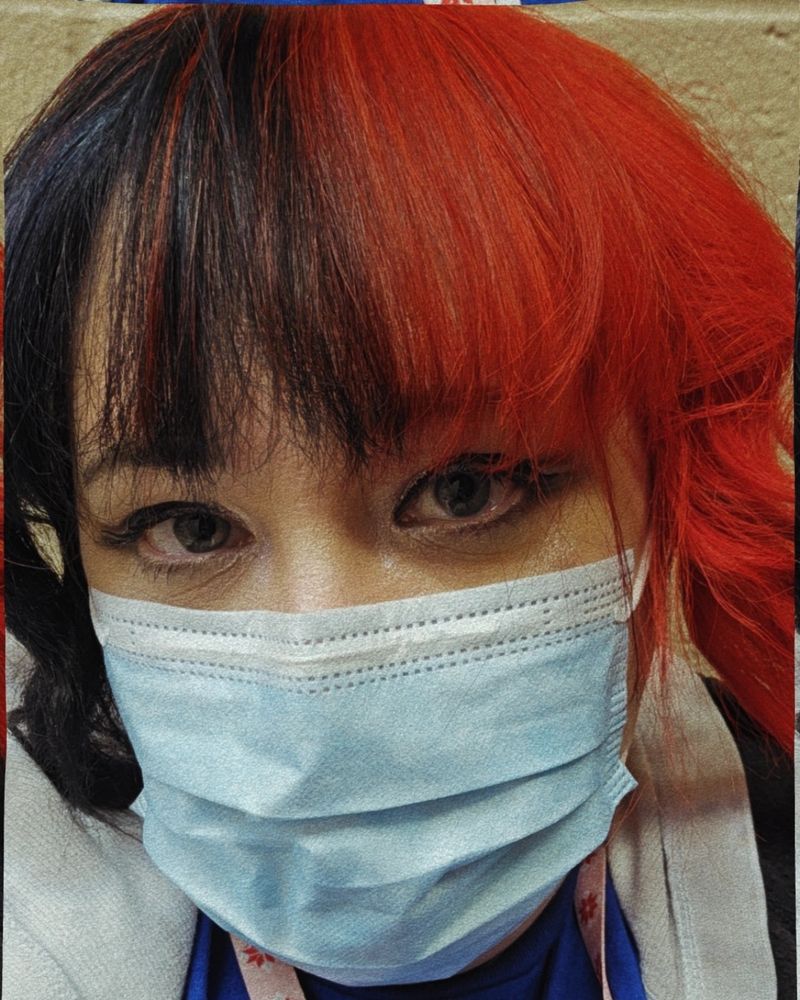 Photo of me. White woman in her 40s. Her hair is split dyed black and fire engine red. She's wearing a blue face mask. 