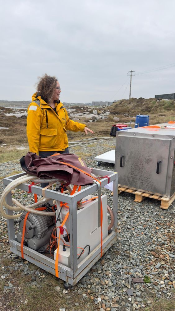 Photo of a woman in a yellow jacket and light brown curly hair explaining something next to a metallic box about half her height. In the fron a rack with sme tubing and a machine and some plastic covers of similar size is placed.