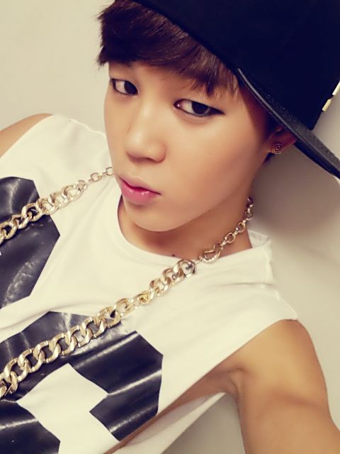 Jimin from BTS sometime close to their debut in 2013, wearing a black baseball cap turned sideways, a white tank top with black numbers on it, a large gold chain, and lots of eyeliner 