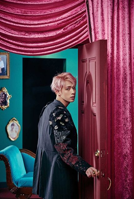Jin of BTS with pink hair, wearing a fancy jacket. He's standing next to an open pink door that leads into a room painted in teal with a teal couch in it and some portraits on the wall. His hand is on the doorknob. The room he's leaving has pink velvet curtains on the wall and hanging over the door.