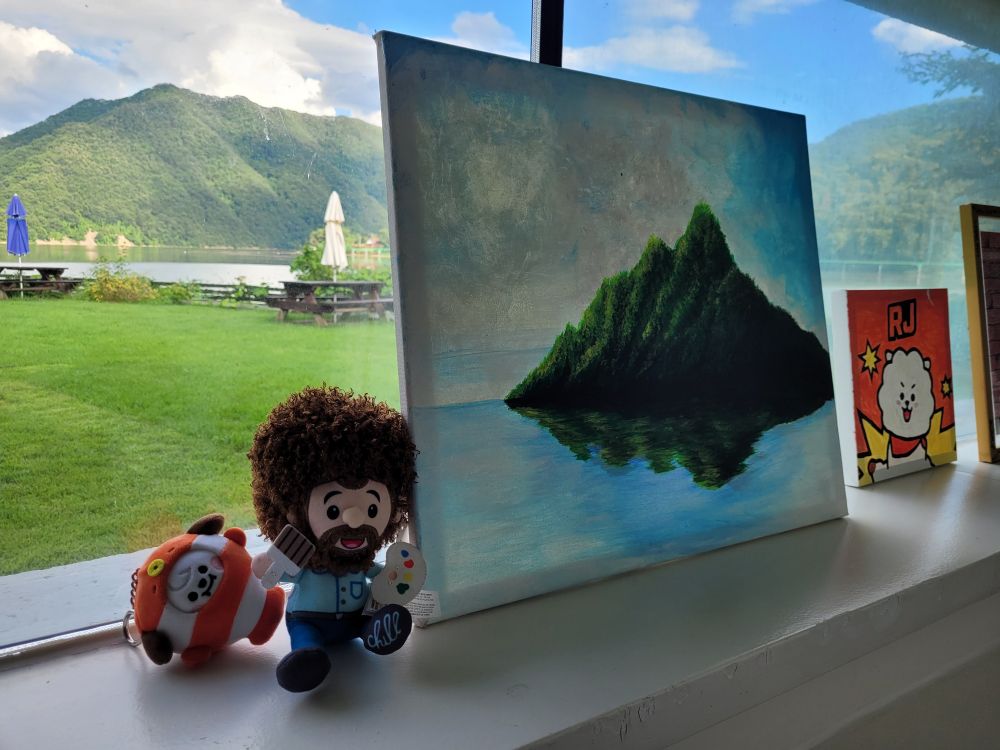 A keychain plushie of BT21 Chimmy in a clownfish costume next to a Bob Ross plushie, a painting of a green mountain over clear blue water, and a painting of RJ of BT21. Behind all these items is a window through which we can see a green lawn, a few picnic tables with umbrellas, a lake, hills covered with green trees, and a blue sky with fluffy white clouds.