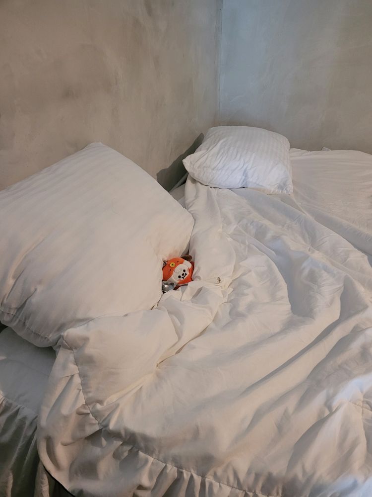 A keychain plushie of BT21 Chimmy in a clownfish costume posed in a bed with a spoon positioned as if he's holding it 