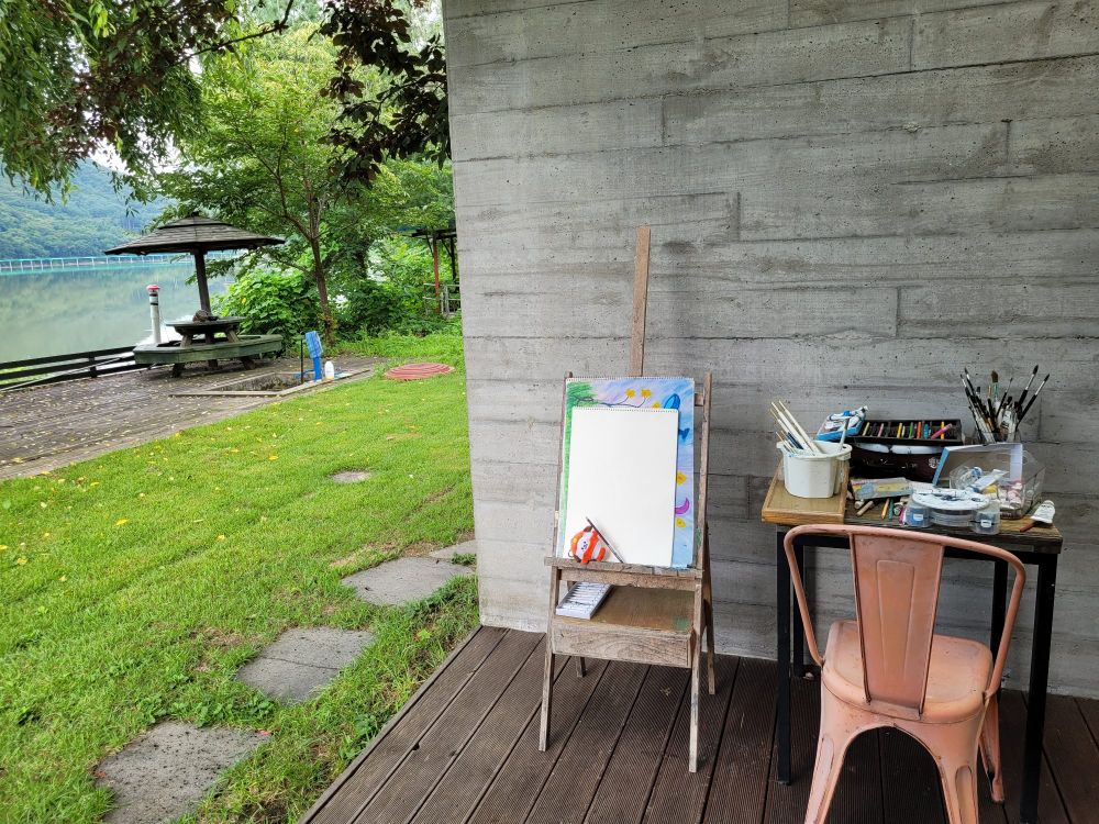 An artist's easel, an orange metal chair, and a table holding paintbrushes, paints, and colored pencils sitting outdoors on a deck, with a concrete wall behind them. To the left are a green lawn with stepping stones, trees, and another deck looking out over water. A keychain plushie of BT21 Chimmy in a clownfish costume is propped up on the easel in front of some paper with a purple colored pencil leaning on him. If you zoom in and look closely, you can see that Chimmy drew a purple heart for you 💜 