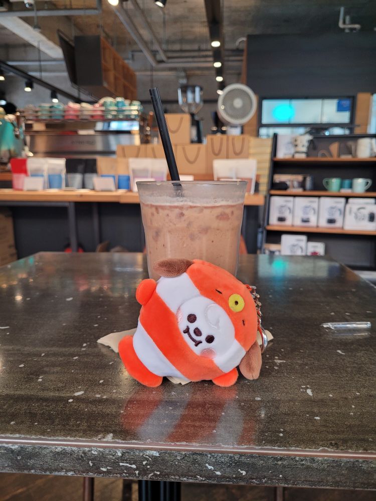 A keychain plushie of BT21 Chimmy in a clownfish costume posed in front of an iced mocha at a coffee shop 