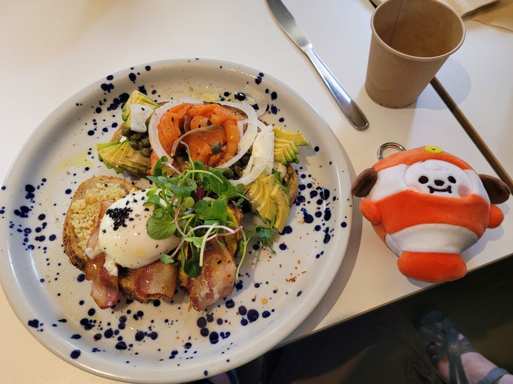 A keychain plushie of BT21 Chimmy in a clownfish costume next to a plate that has two pieces of toast, one topped with bacon and an over easy egg, the other with smoked salmon, onions, capers, and avocado slices. The farmer's plate at the Analog Garden cafe is very tasty, it turns out.